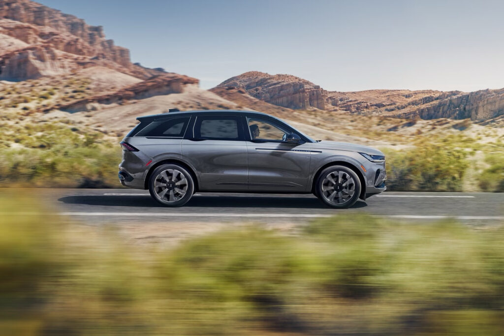 Gray SUV driving on desert highway with rocky hills in the background, showcasing speed and adventure.