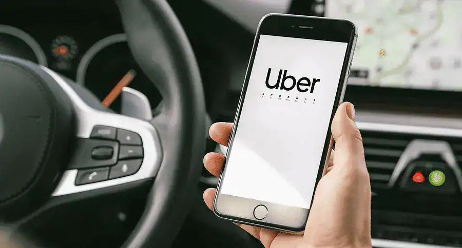 Person holding a smartphone displaying the Uber app inside a car, with the steering wheel and dashboard visible.