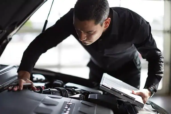 Mechanic inspecting a car engine with a clipboard under a raised hood, in a garage setting.