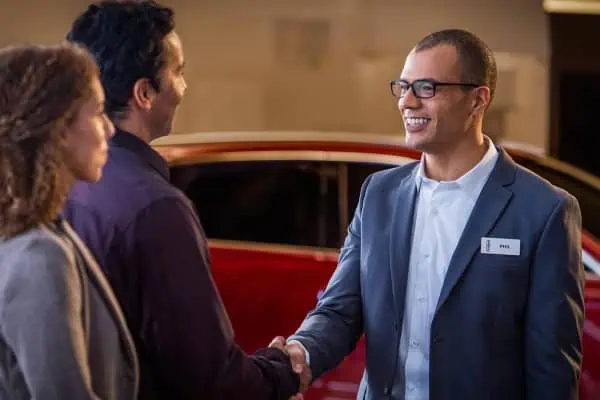 Salesperson with glasses shakes hands with a smiling couple in front of a red car.
