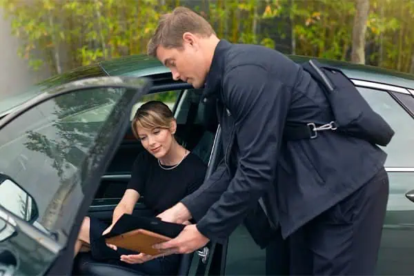 A chauffeur hands a tablet to a woman seated in a car, with lush greenery in the background.