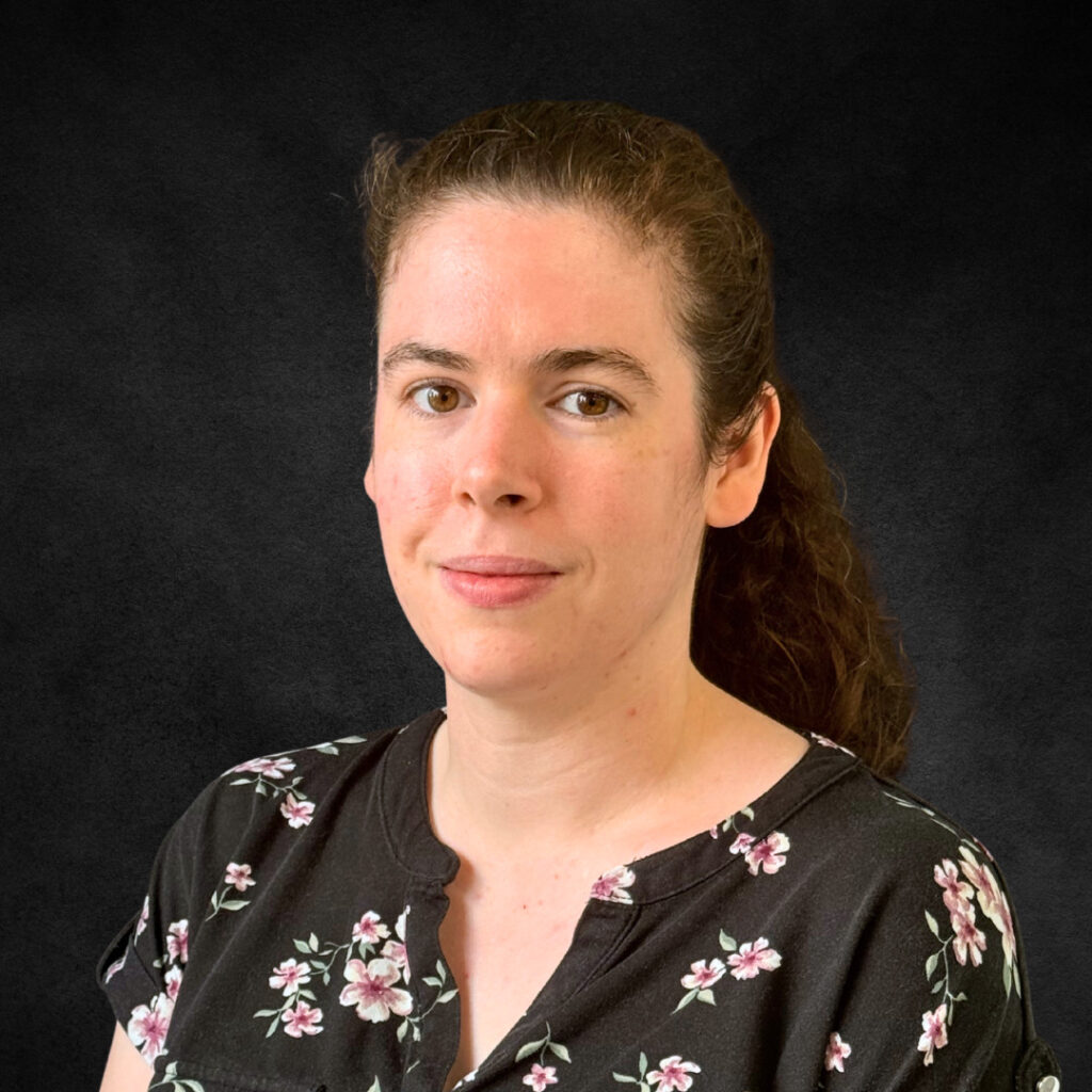 Smiling woman with dark hair in a ponytail wearing a floral blouse on a dark background.